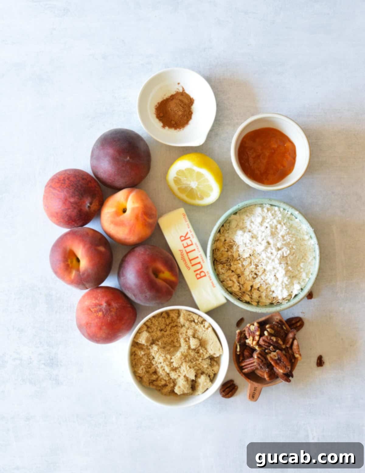 A flat lay of key ingredients for peach crisp: fresh peaches, lemon, peach preserves, flour, rolled oats, pecans, unsalted butter, and brown sugar.