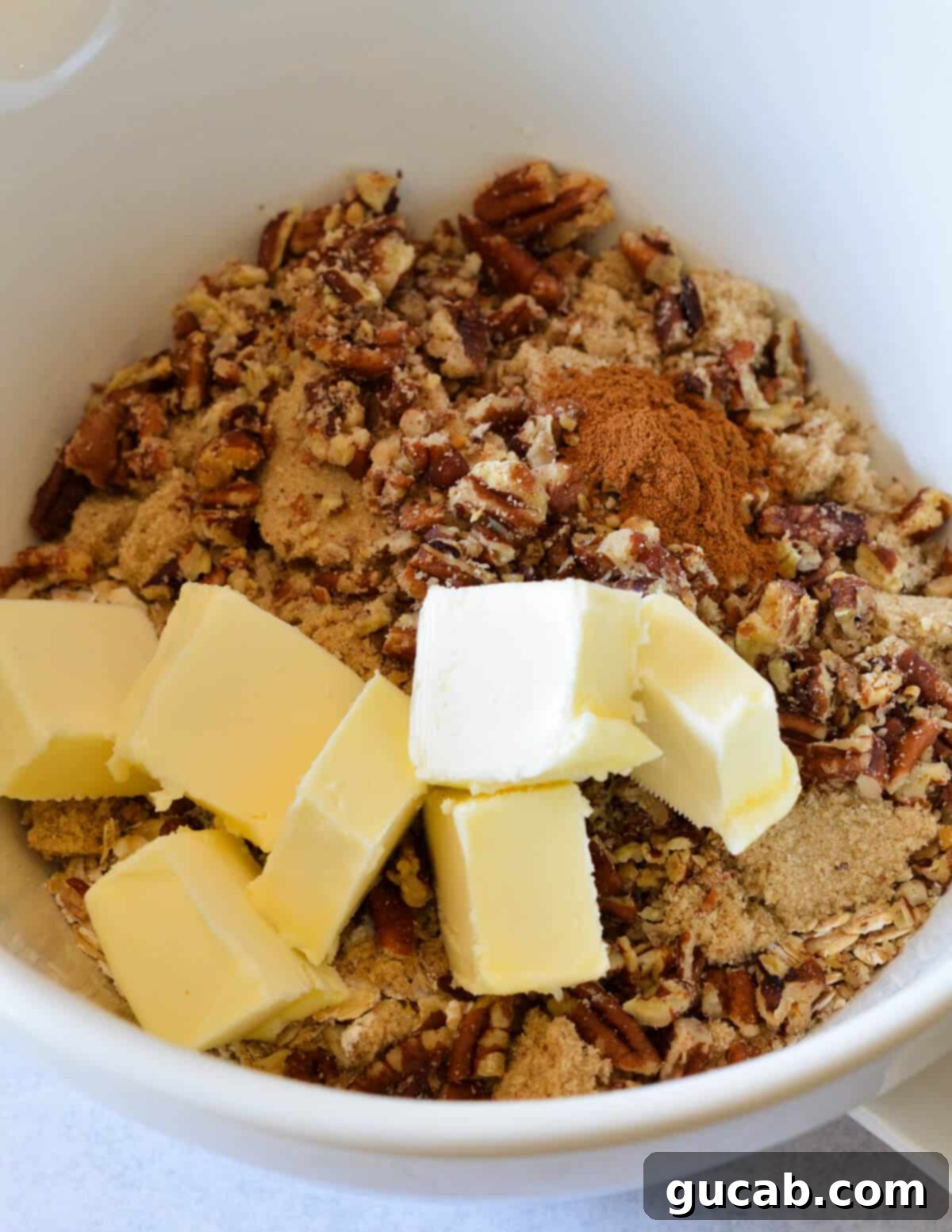 A bowl containing raw crumble topping ingredients: oats, brown sugar, cinnamon, crushed pecans, flour, and cold butter pieces, ready to be mixed.