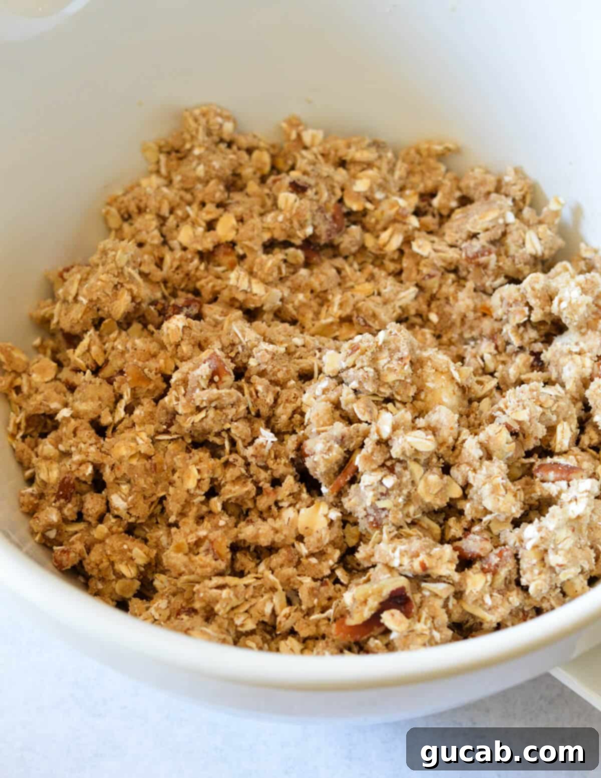 A bowl of fully prepared oat crumble topping, showing a coarse, sandy texture with visible oat and pecan pieces.