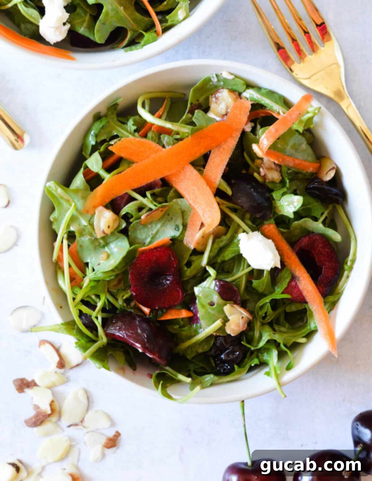 A vibrant bowl of arugula salad brimming with fresh red cherries, delicate shaved carrots, creamy feta cheese crumbles, and glistening candied almonds, ready to be tossed and enjoyed.