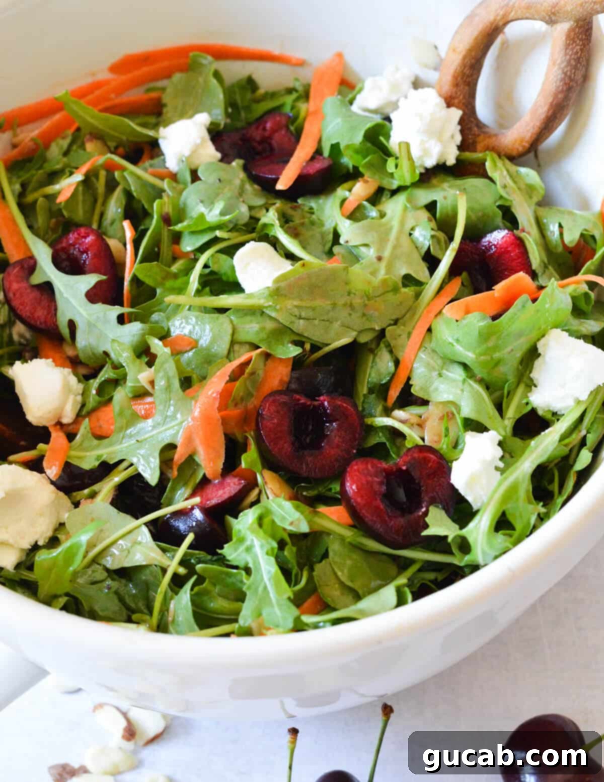 A close-up of a beautifully plated arugula salad with cherries, shaved carrot ribbons, creamy goat cheese, and a light balsamic vinaigrette dressing, showcasing its fresh ingredients.
