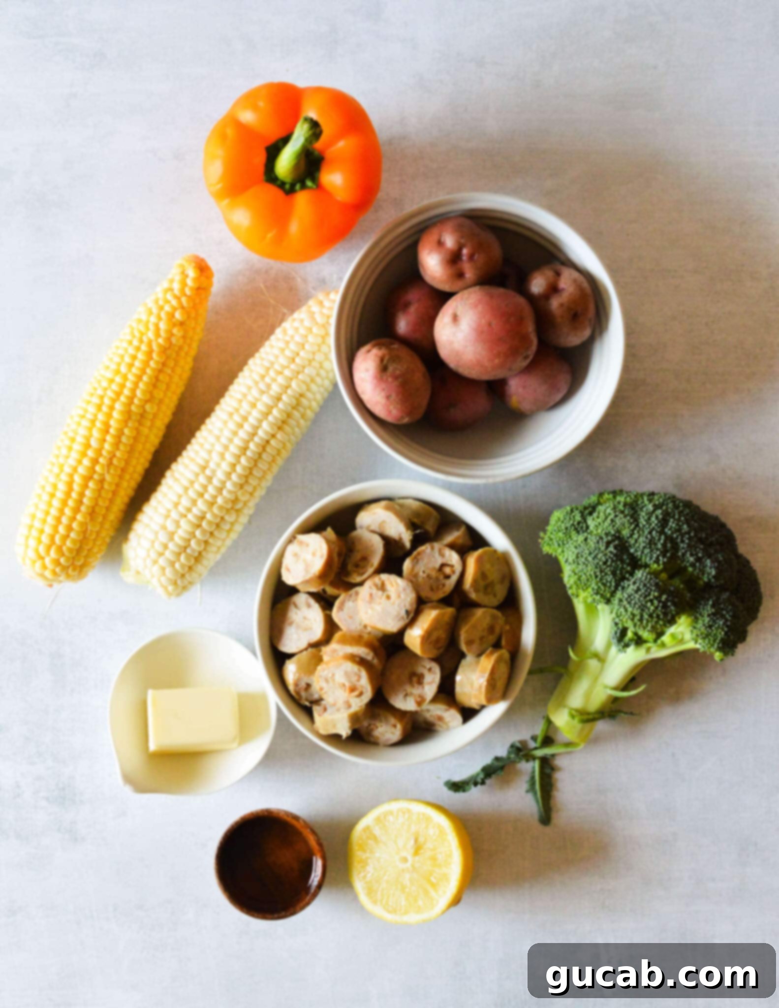 A vibrant selection of fresh ingredients laid out, including colorful bell peppers, small red potatoes, corn on the cob, raw chicken sausage links, fresh broccoli florets, a stick of butter, a bottle of olive oil, and a bright lemon, essential for the sheet pan dinner.