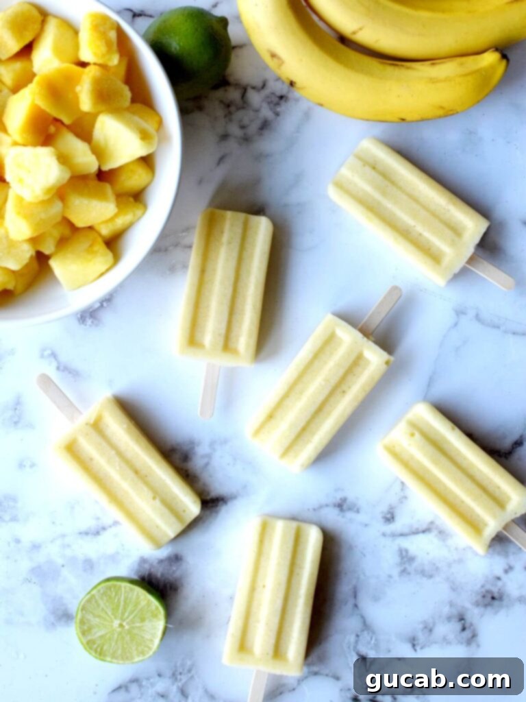 All Fruit Pineapple Popsicles 2 pineapple popsicles laying on a counter with limes, bananas and pineapple around it