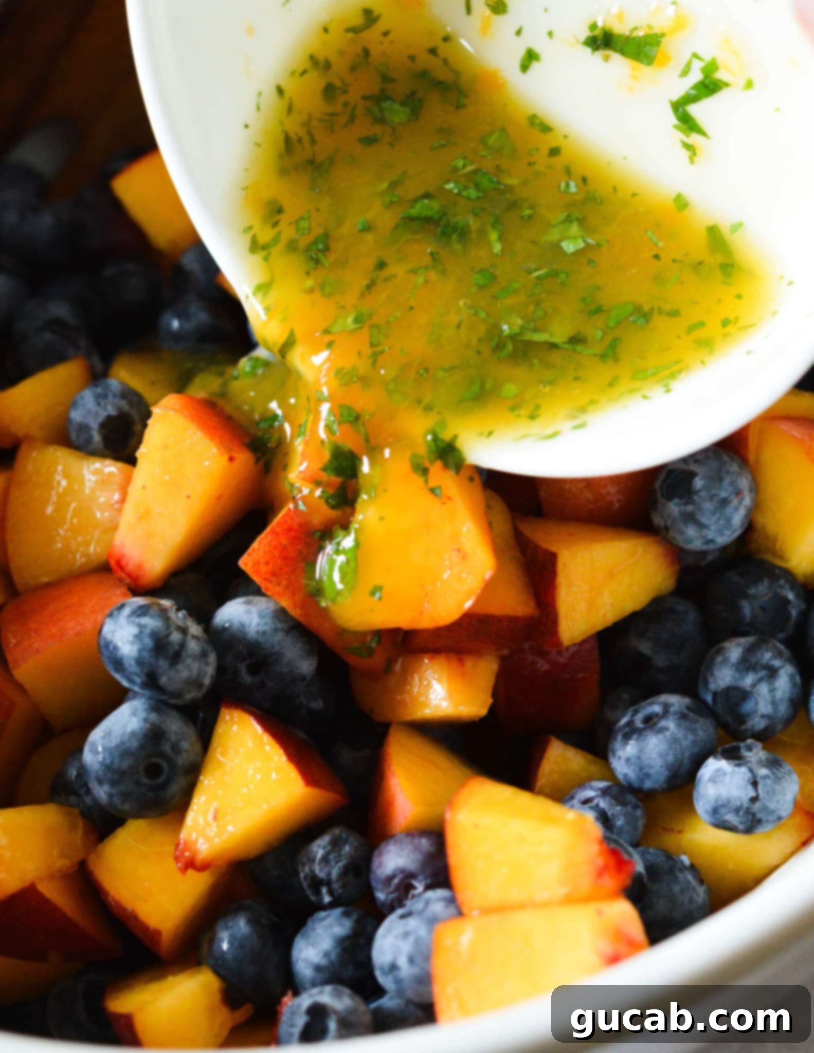 A close-up of the citrus-mint dressing being poured over a bowl of fresh peaches and blueberries, ready to be tossed.
