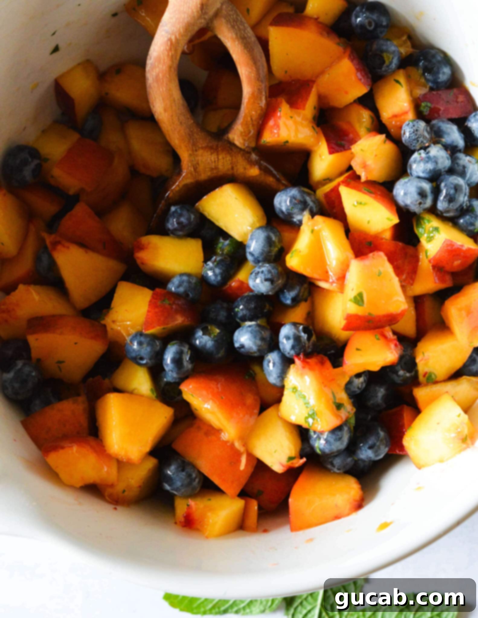 A large bowl of peach and blueberry fruit salad, with a wooden spoon resting inside, ready for serving. 
