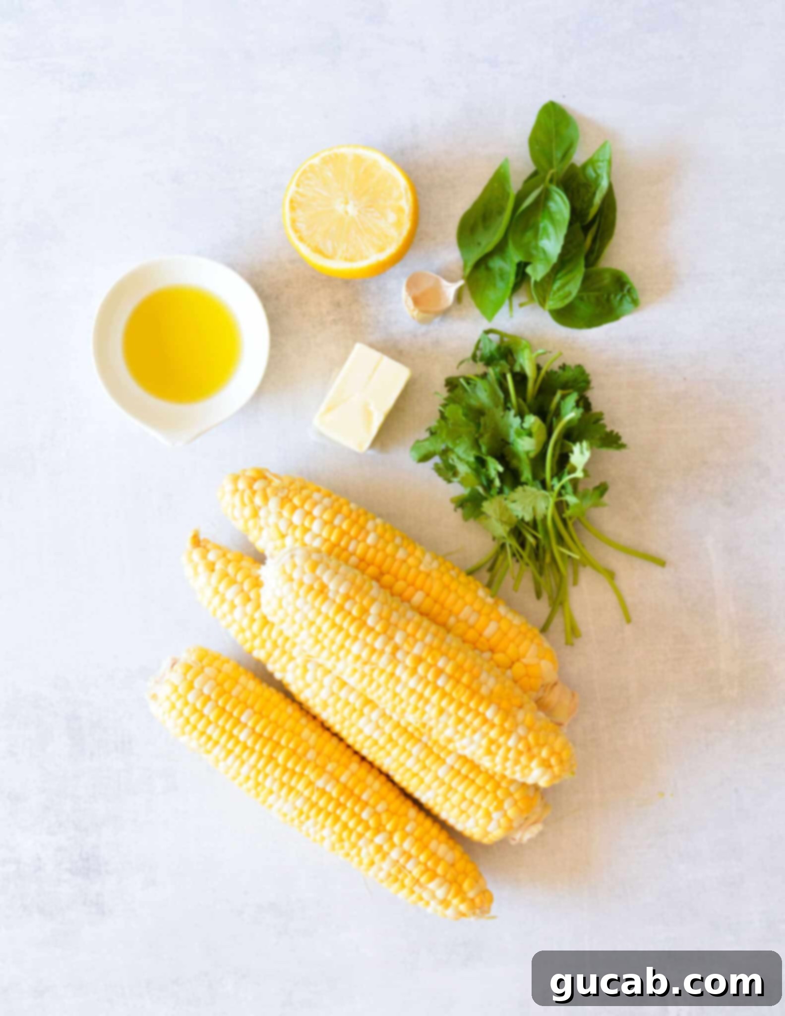 Fresh ears of corn, vibrant green cilantro and basil, a stick of butter, a bright yellow lemon, and a clove of garlic arranged on a surface, highlighting the key ingredients for the recipe.