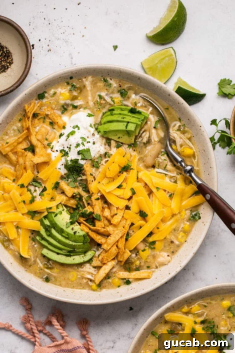 A bowl of creamy white chicken chili, generously topped with shredded cheese, avocado slices, and crispy tortilla strips.
