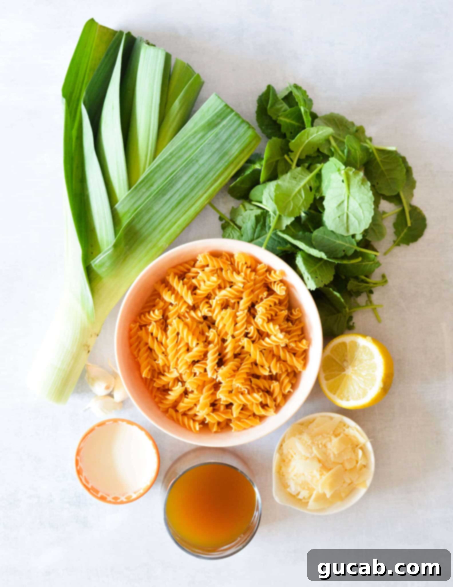 A vibrant flat lay of all the essential ingredients for creamy kale pasta: uncooked pasta, fresh baby kale, sliced leeks, whole garlic cloves, a bright lemon, heavy cream, shaved parmesan, and vegetable broth.