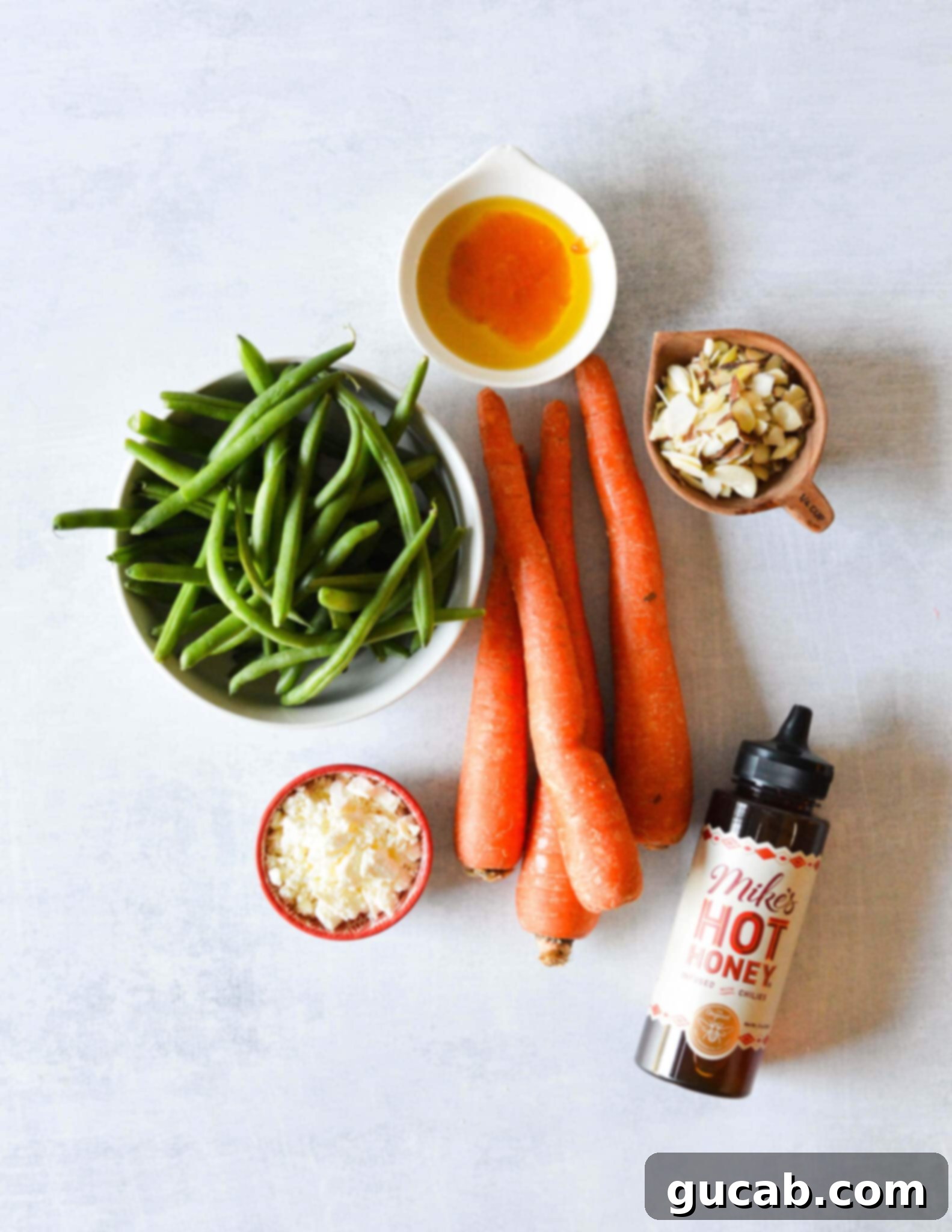 A selection of fresh ingredients laid out: crisp green beans, large carrots, golden olive oil, sweet peach jam, spicy hot honey, thinly sliced almonds, and crumbly feta cheese.