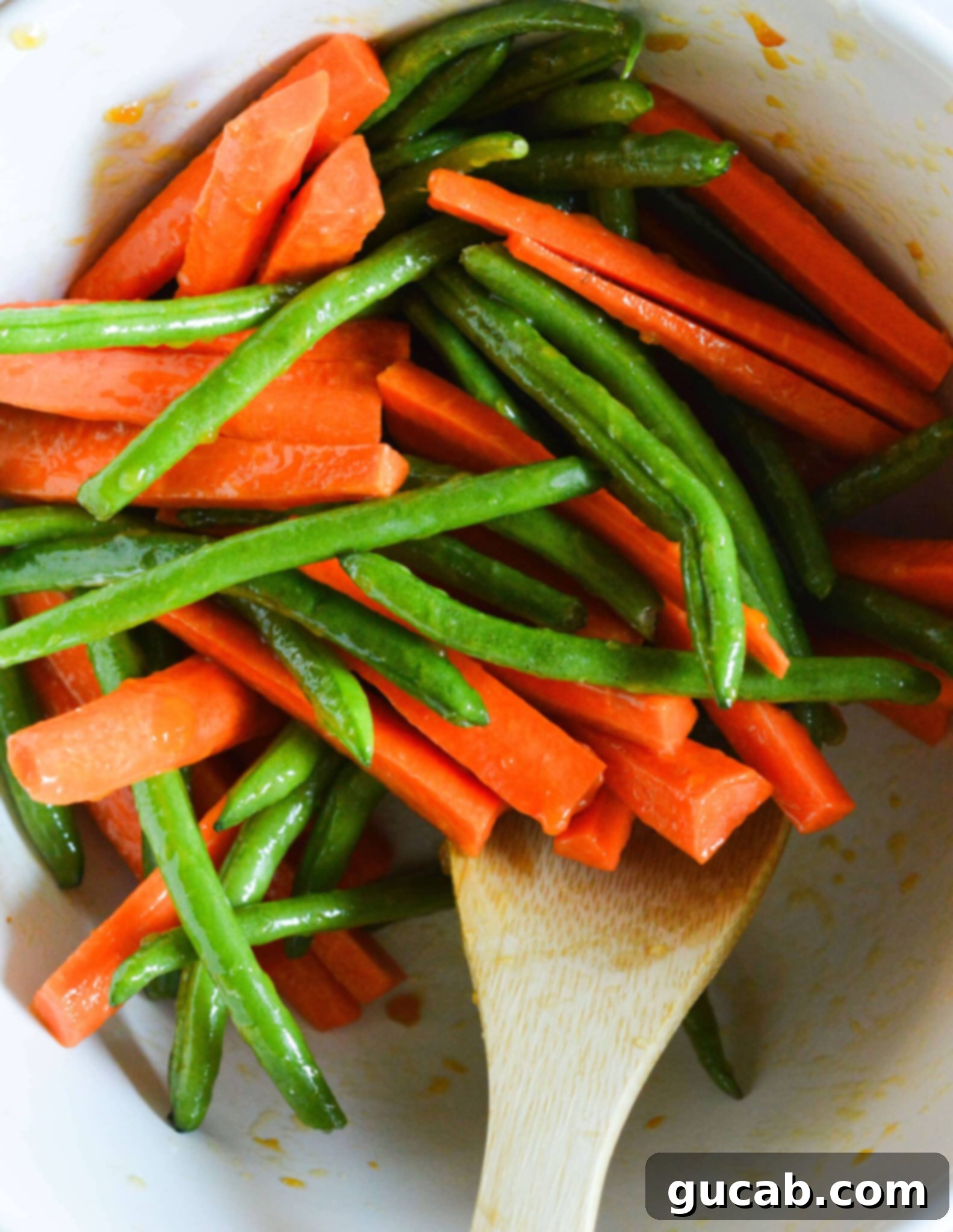 A bowl filled with fresh green beans and evenly sliced carrots, mixed together with a wooden spoon, ready for seasoning.