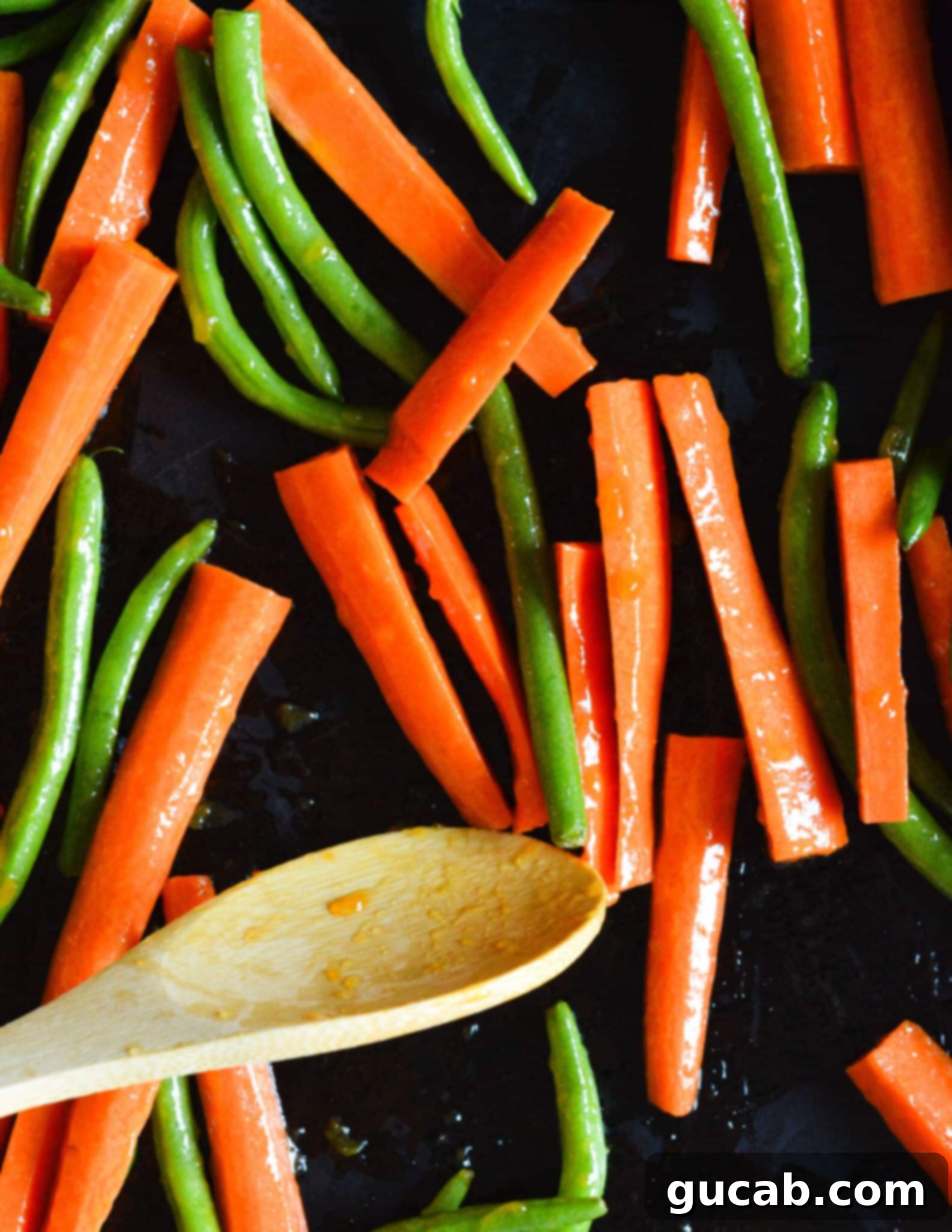 Fresh green beans and sliced carrots coated in glistening olive oil and seasoning, spread out in a single layer on a baking sheet, ready for the oven.