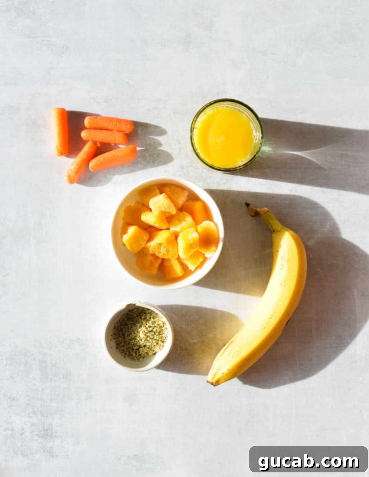 A flat lay showing fresh bananas, vibrant baby carrots, bright orange juice, frozen mango chunks, and a small bowl of hemp hearts, ready for blending into a delicious smoothie.