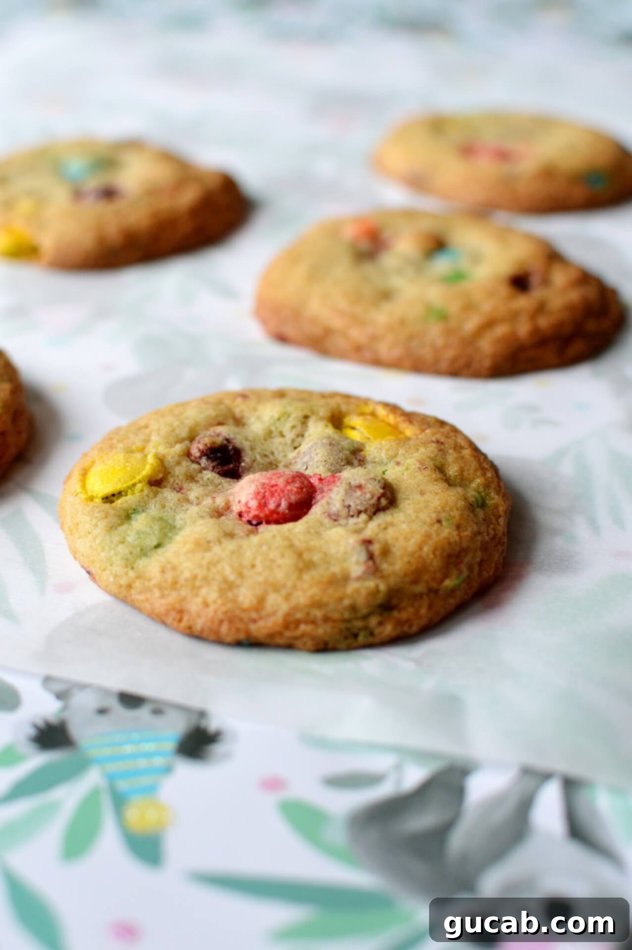 Five perfectly round gluten-free M&M cookies resting on a sheet of parchment paper, showcasing their even spread and colorful candies.