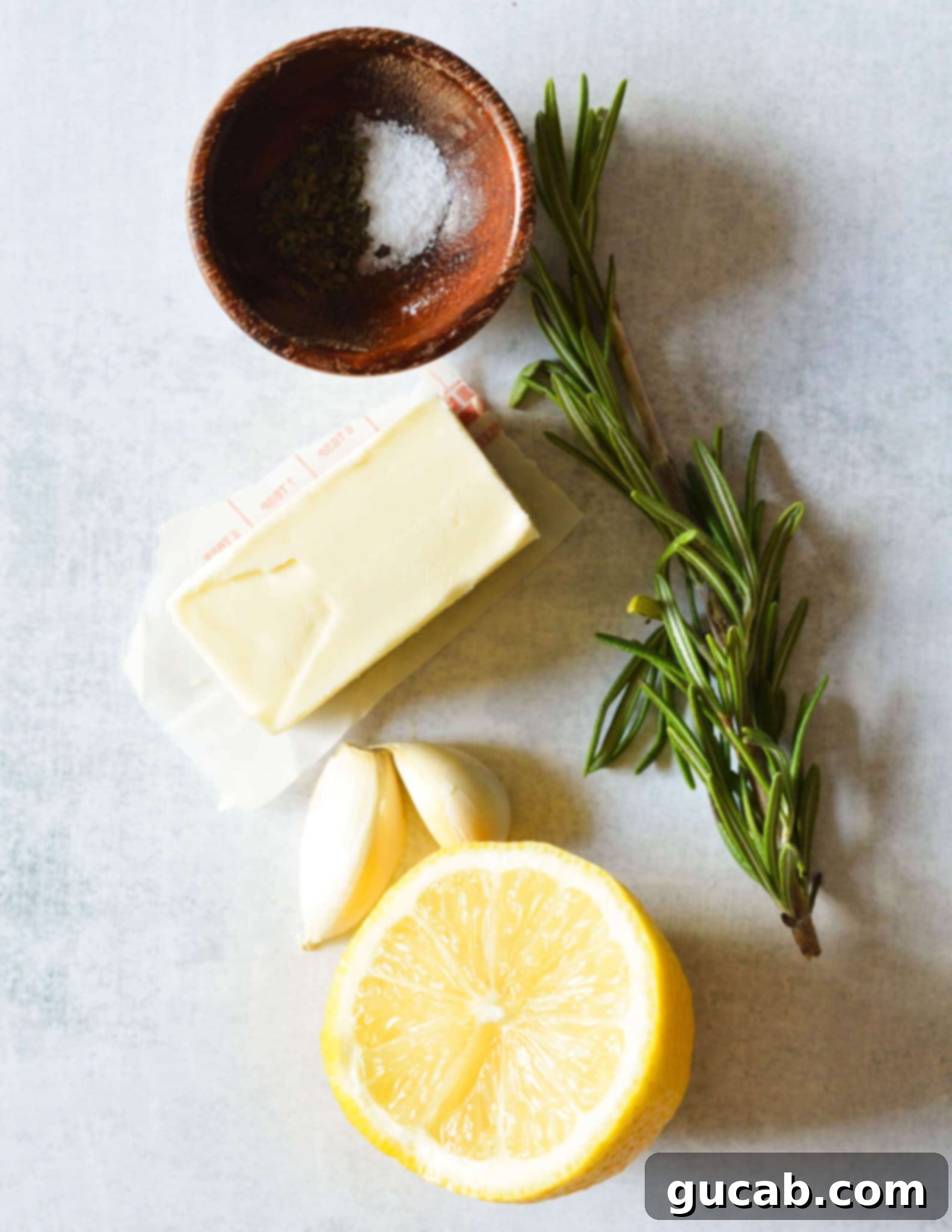 A flat lay of the key ingredients for rosemary garlic butter: a lemon, fresh garlic cloves, fresh rosemary sprigs, a block of butter, and a small pile of salt.