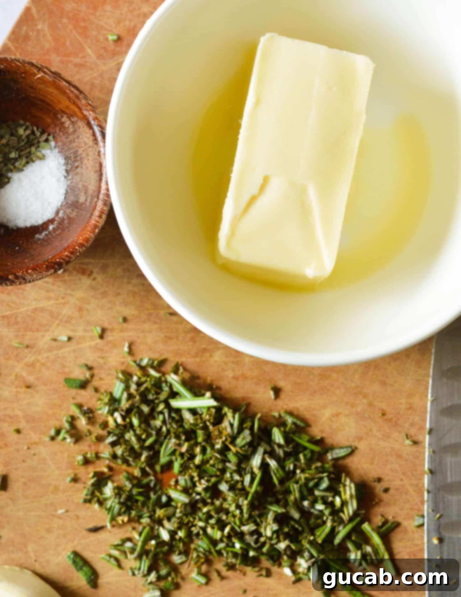 A bowl with softened butter, next to it minced fresh rosemary on a cutting board.