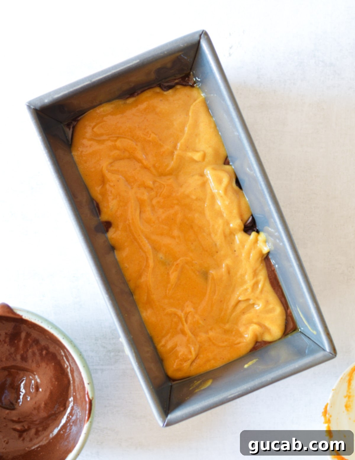 A layer of pumpkin batter spread over the chocolate layer in the loaf pan, showing the start of the marble effect.