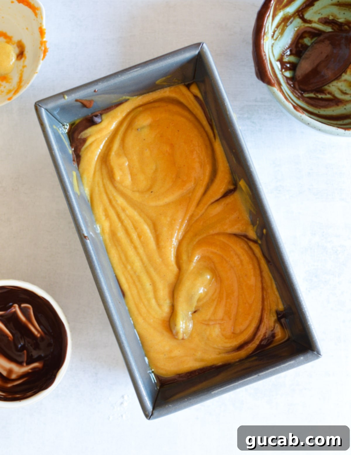 A loaf pan filled with swirled pumpkin and chocolate batter, showing the intricate marble pattern before baking.