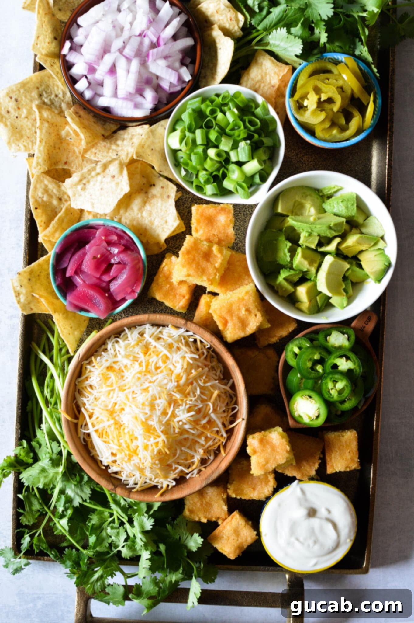 A vibrant chili bar spread featuring bowls of diced red onion, fresh cilantro, pickled jalapenos, sliced green onions, pickled red onions, creamy diced avocado, crunchy tortilla chips, fluffy cornbread squares, shredded cheese, and rich sour cream, all ready for guests to customize their perfect bowl.