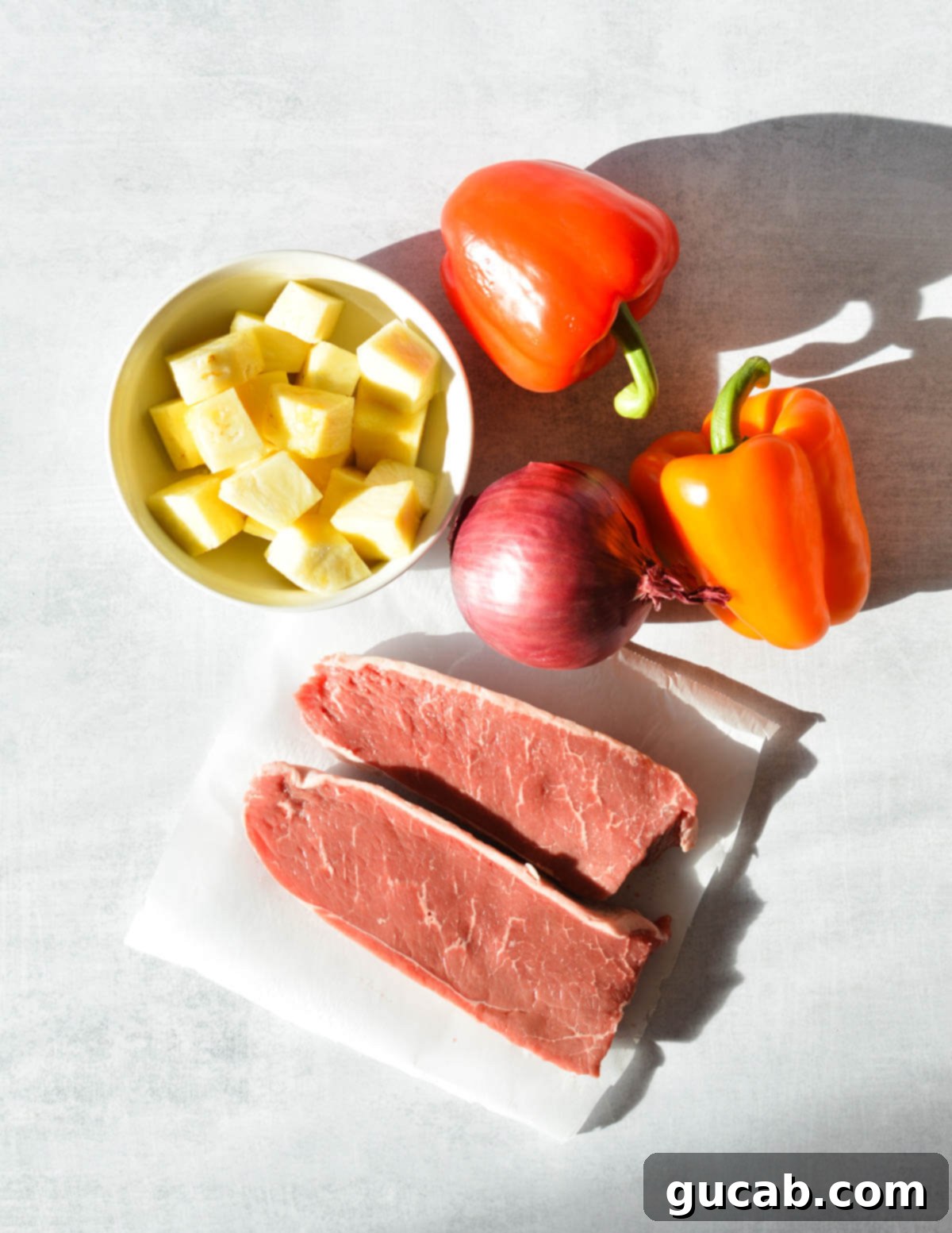 Raw ingredients for steak kebabs: bell peppers, fresh pineapple chunks, sirloin steak cubes, and sliced red onion on a wooden board.