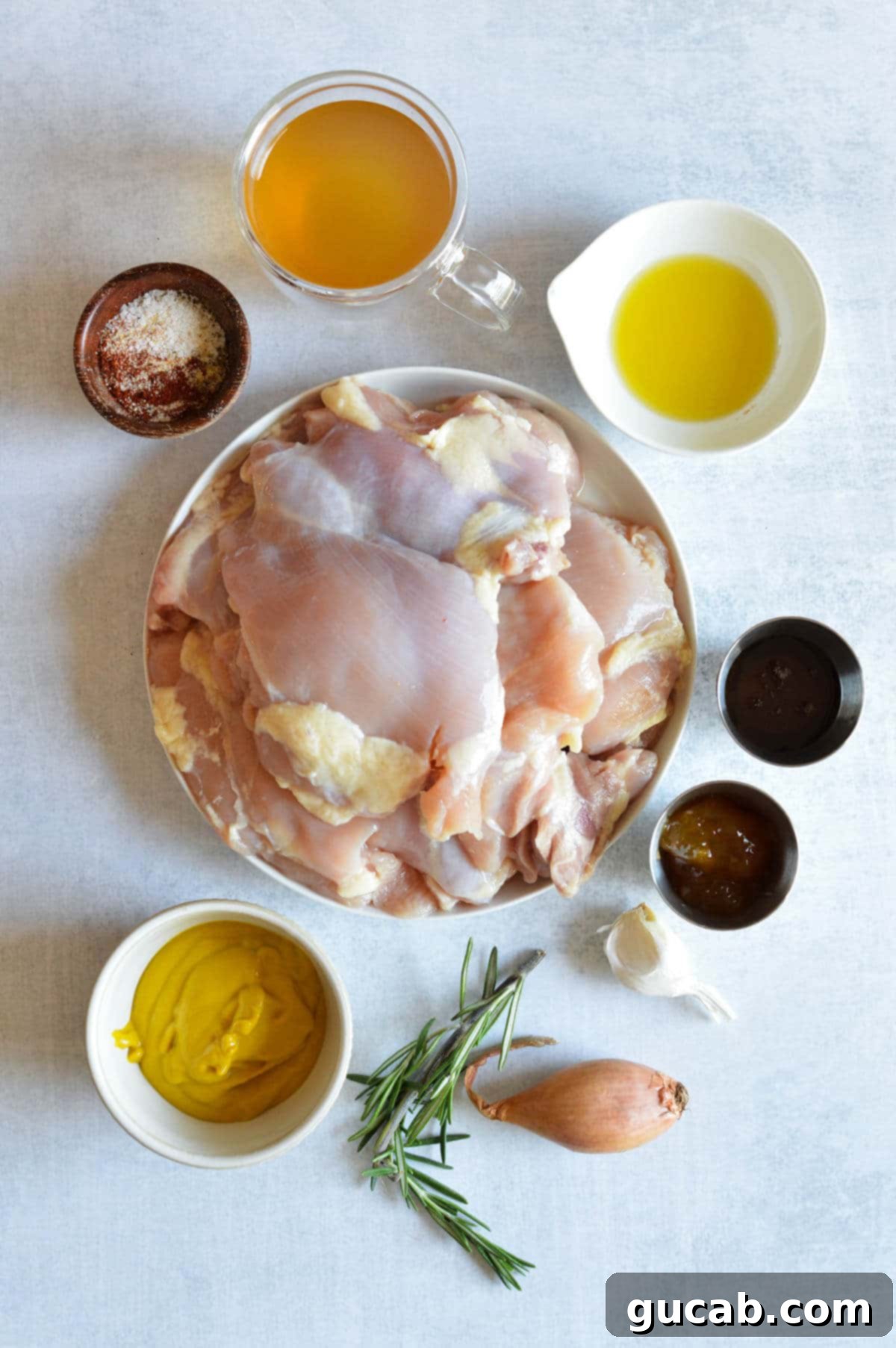 A flat lay photograph of all key ingredients for mustard chicken: boneless skinless chicken thighs, olive oil, a mix of spices, chicken stock, yellow mustard, apricot jam, honey, minced garlic, thinly sliced shallot, and fresh rosemary sprigs.