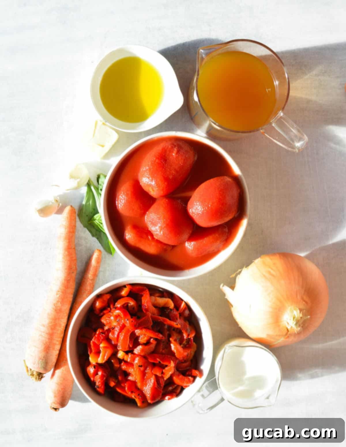 Arrangement of key ingredients: vegetable stock, olive oil, canned tomatoes, onion, roasted red peppers, fresh basil, and carrots.
