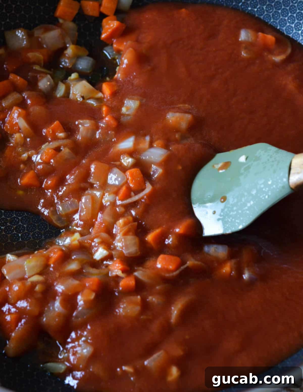 spatula stirring tomato sauce with sauteed carrots and onions in a skillet.