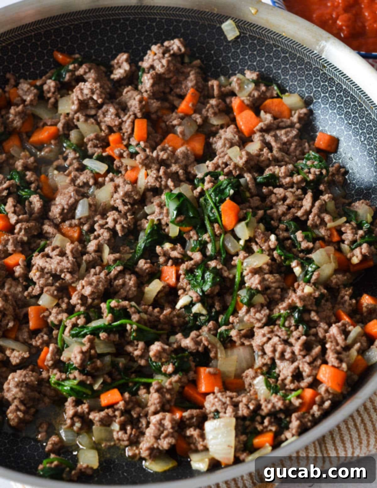 Cooked ground beef in a skillet with spinach and carrots, forming a robust base for the sauce.