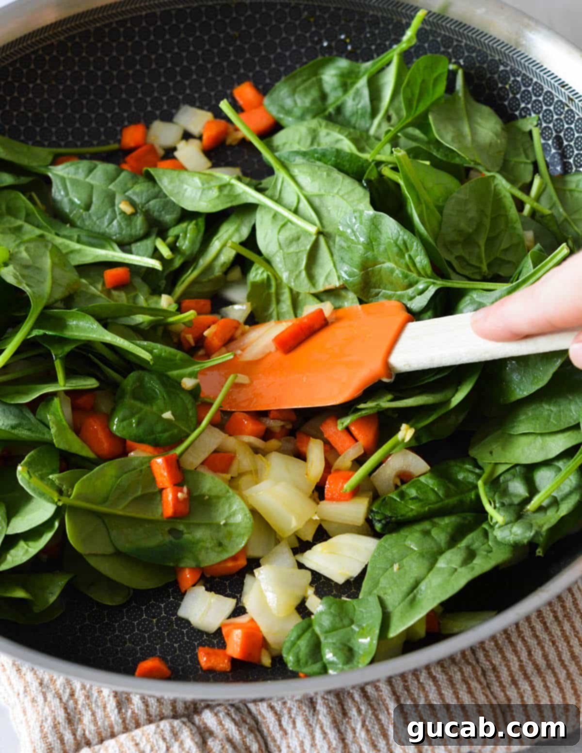 Cooked onions, garlic, and carrots in a skillet with baby spinach on top, ready to be wilted.