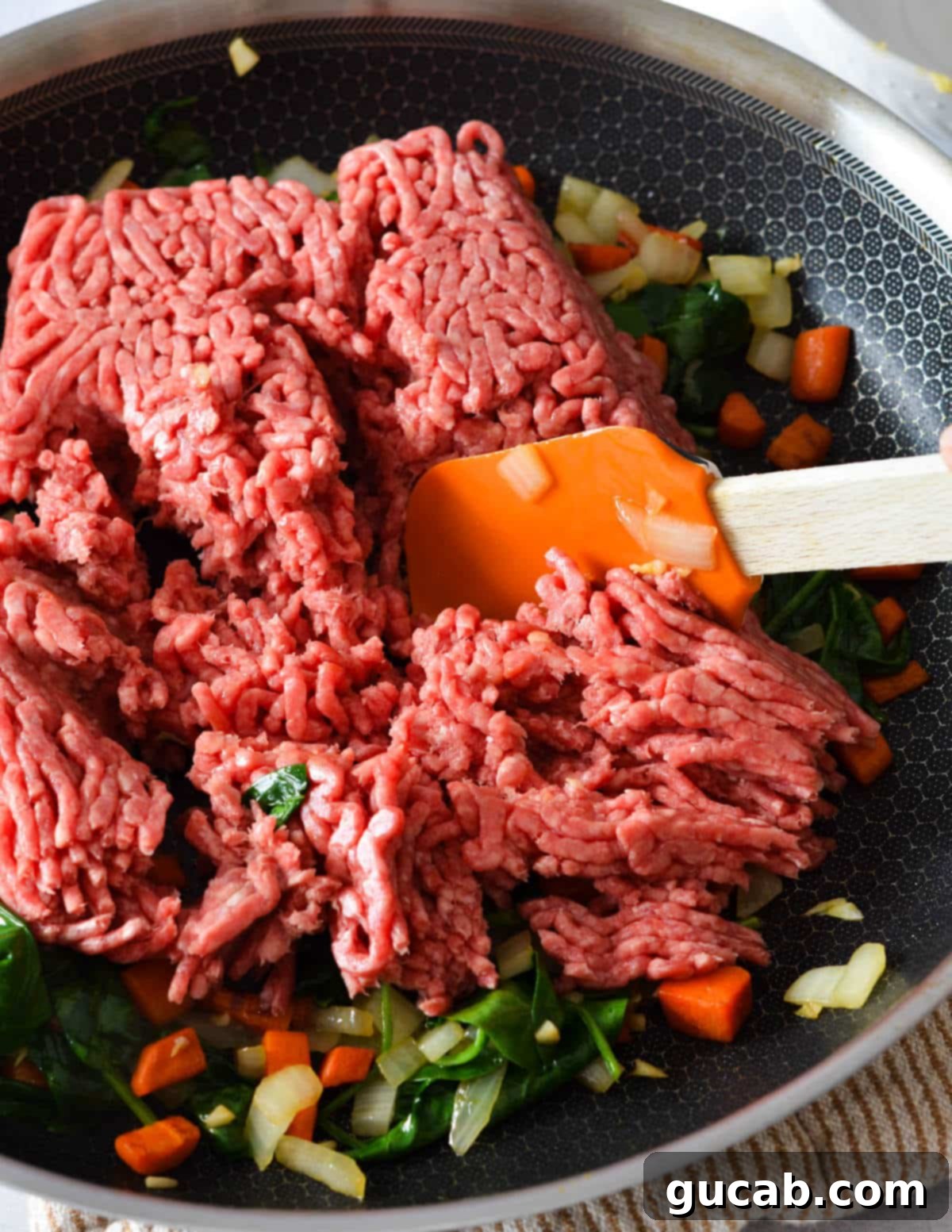 Raw ground beef added to a skillet with sautéed veggies, ready to be cooked.