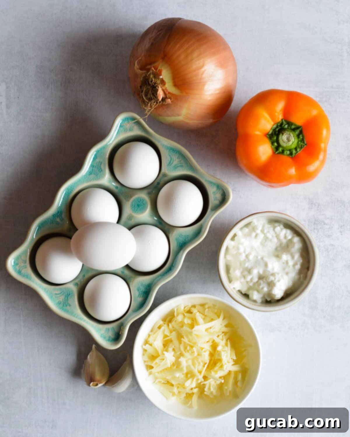 A flat lay of fresh ingredients: white onion, colorful bell pepper, large eggs, block of white cheddar cheese, a tub of cottage cheese, and garlic cloves, ready for making egg muffins.