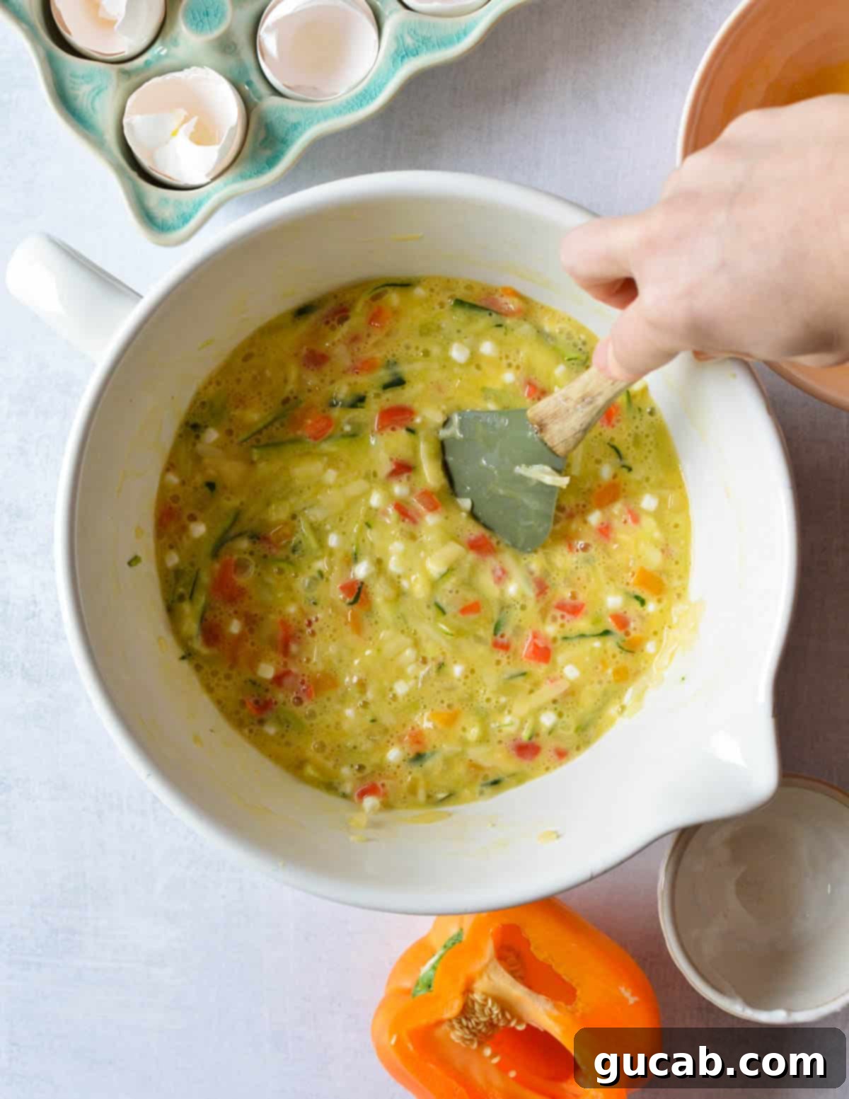 A hand stirring a large bowl filled with a mixture of eggs, cheese, sautéed vegetables, and cottage cheese, ensuring all ingredients are well combined.