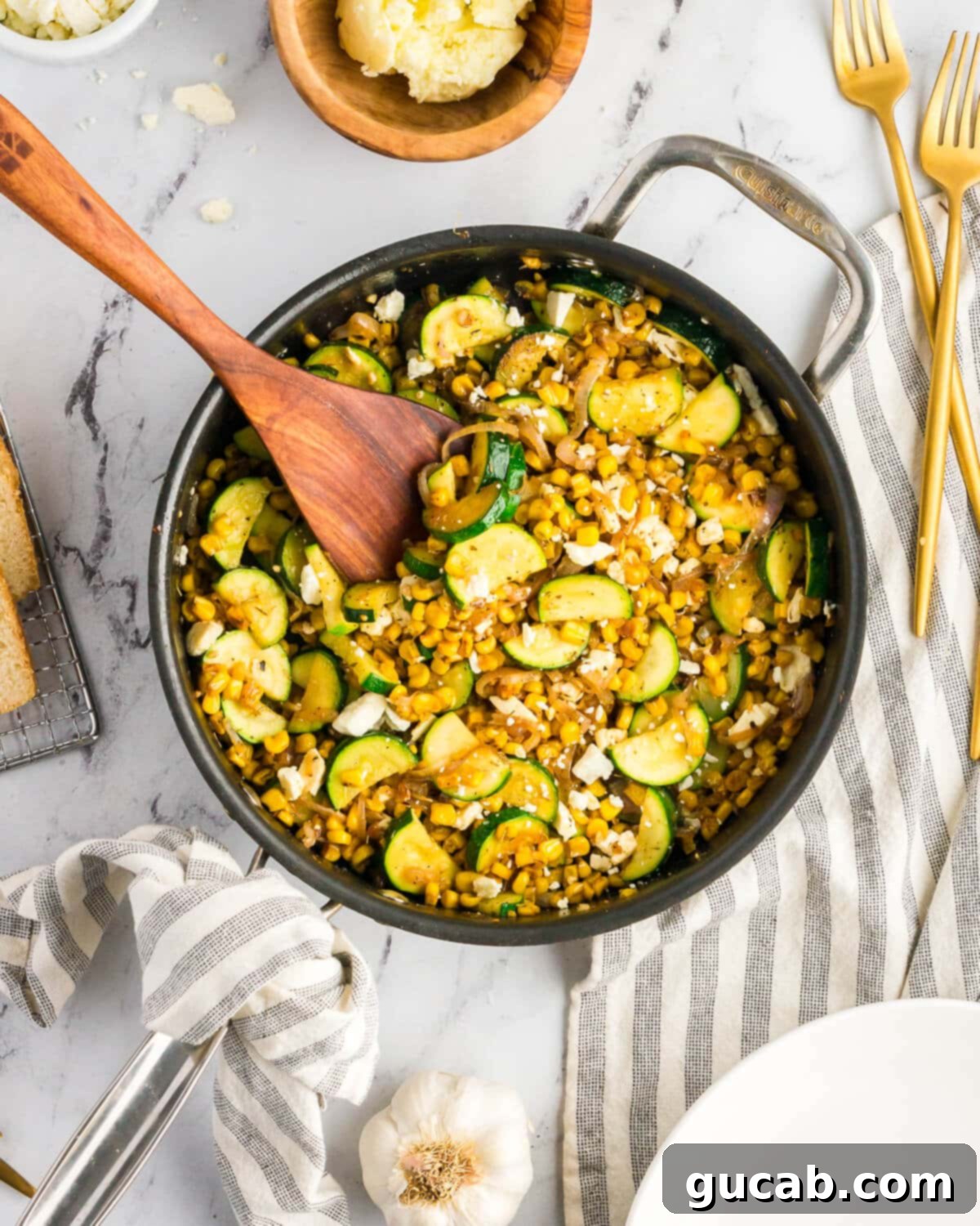 A large skillet filled with the finished dish: perfectly sauteed zucchini, caramelized corn, shallots, and melted feta, with a wooden spoon nestled in it.