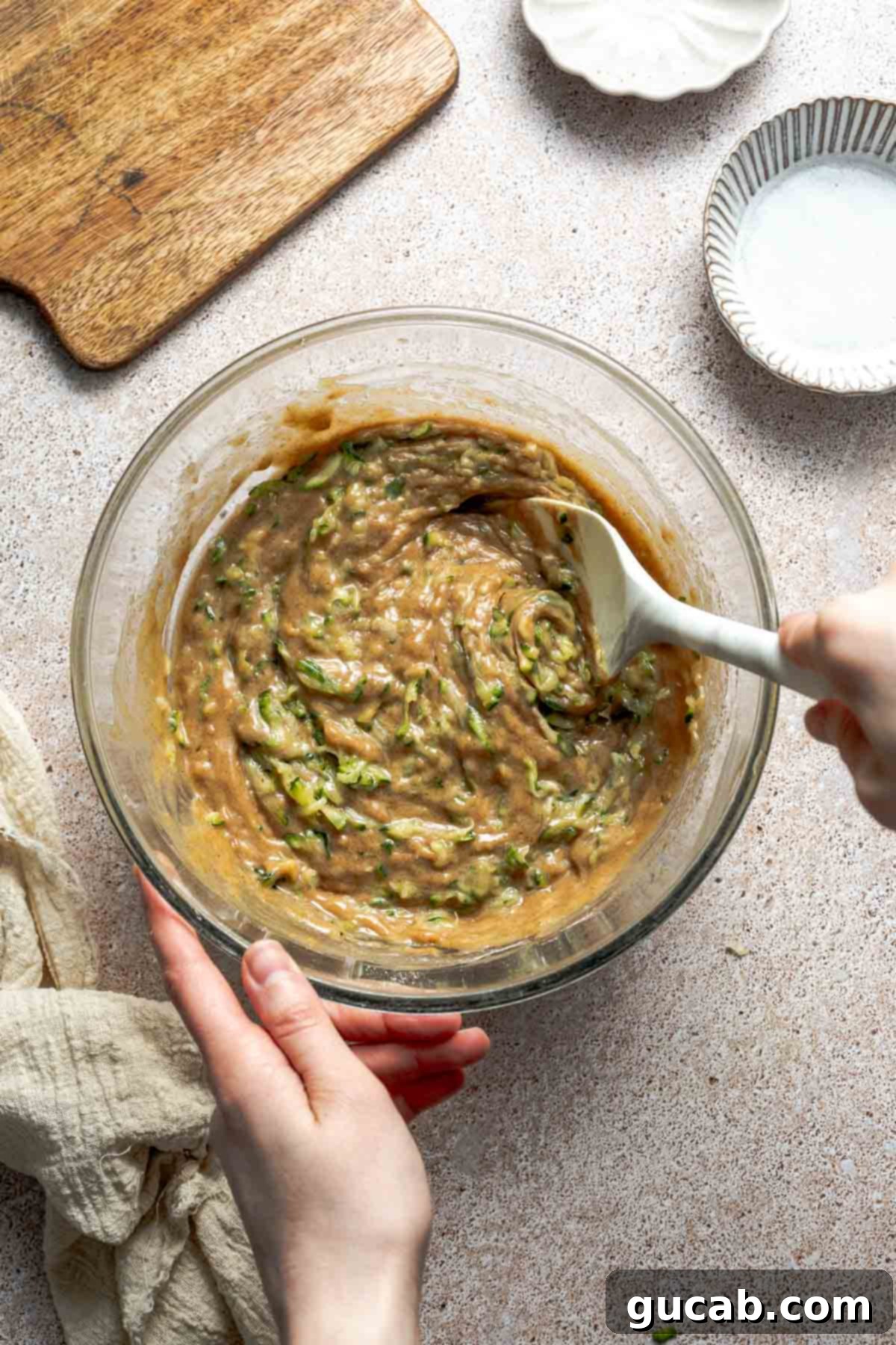 The process of stirring freshly shredded zucchini into the gluten-free bread batter in a mixing bowl using a spatula, ensuring even distribution.