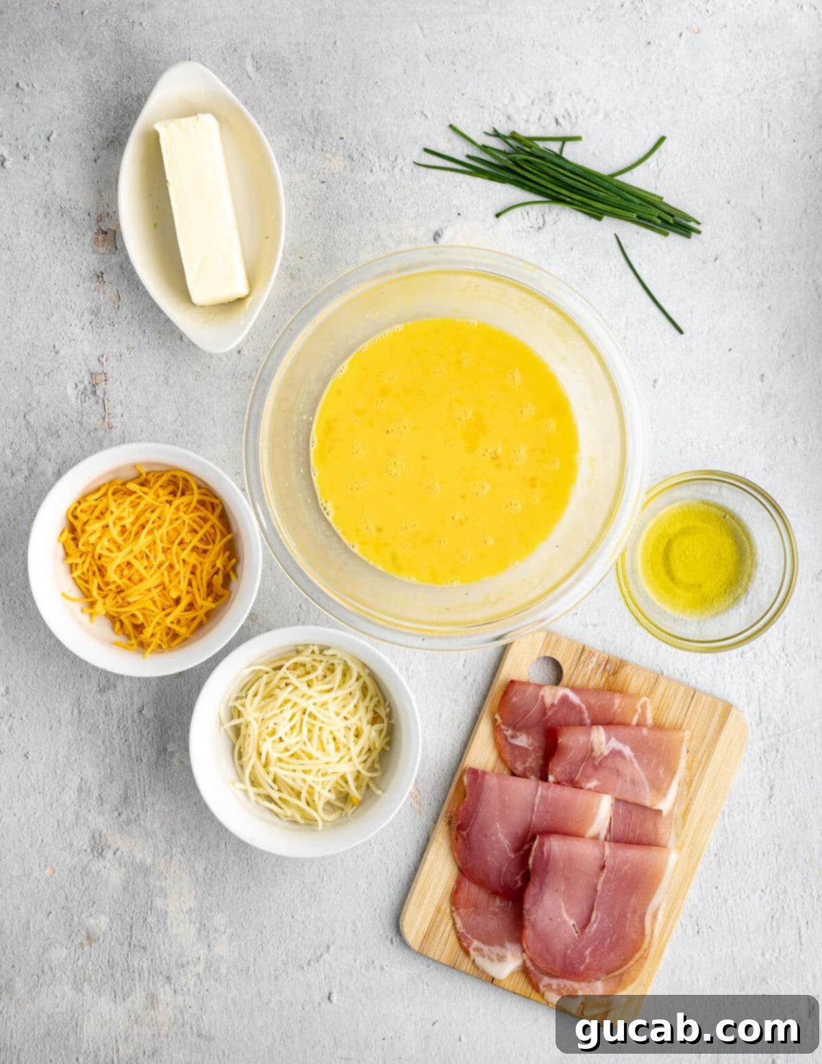 Savory Prosciutto & Cheese Scramble 3 A flat lay photograph showcasing all the ingredients for cheesy scrambled eggs: butter, fresh chives, eggs, blocks of cheddar and gruyere cheese, olive oil, and slices of prosciutto.