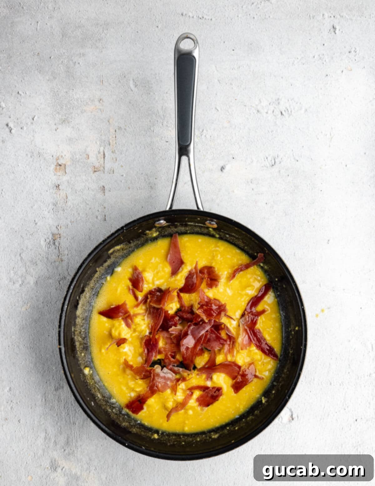 Savory Prosciutto & Cheese Scramble 10 Pieces of torn crispy prosciutto being added over partially cooked scrambled eggs in a skillet.