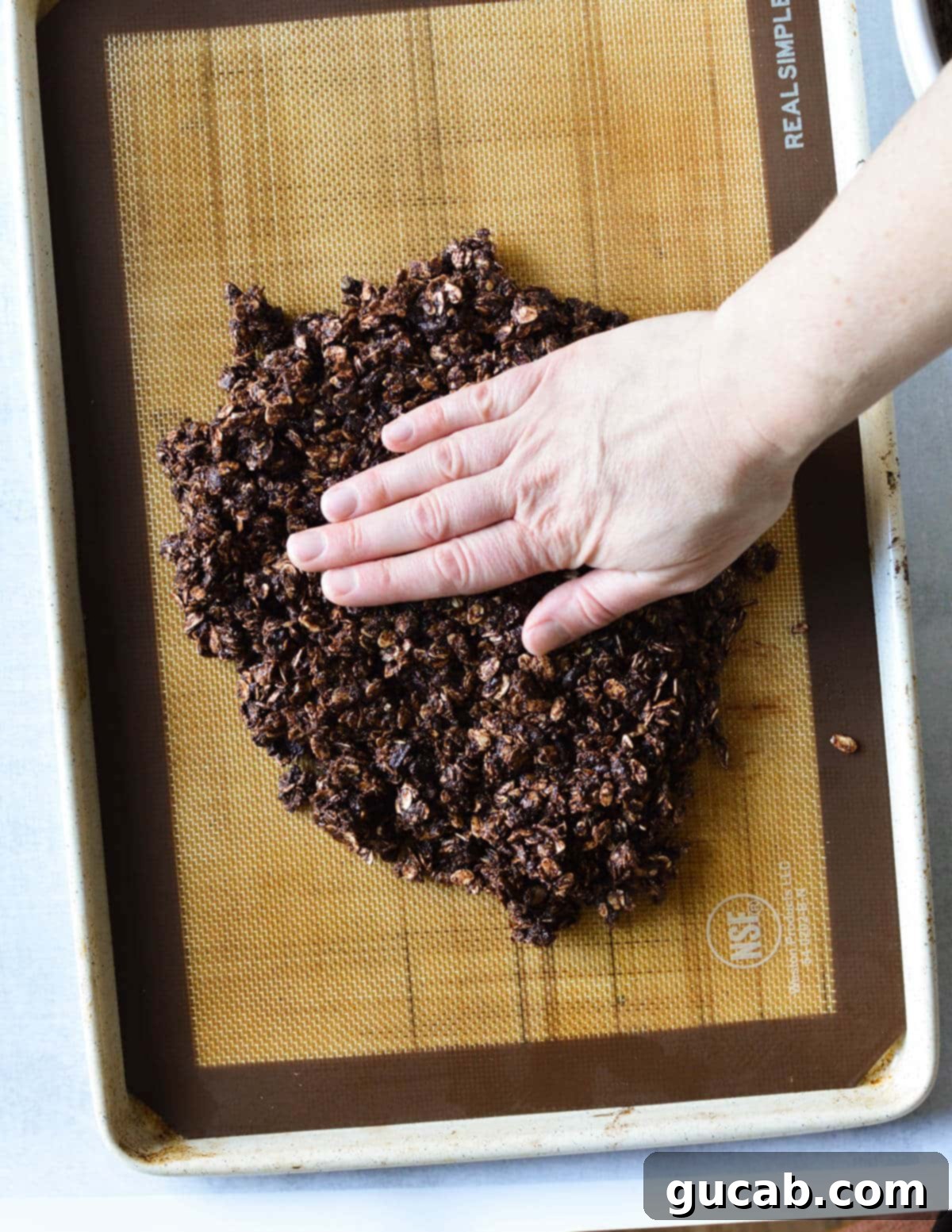 Dark Chocolate Granola 12 A hand firmly pressing the chocolate granola mixture down onto the sheet pan, forming an even layer for baking.