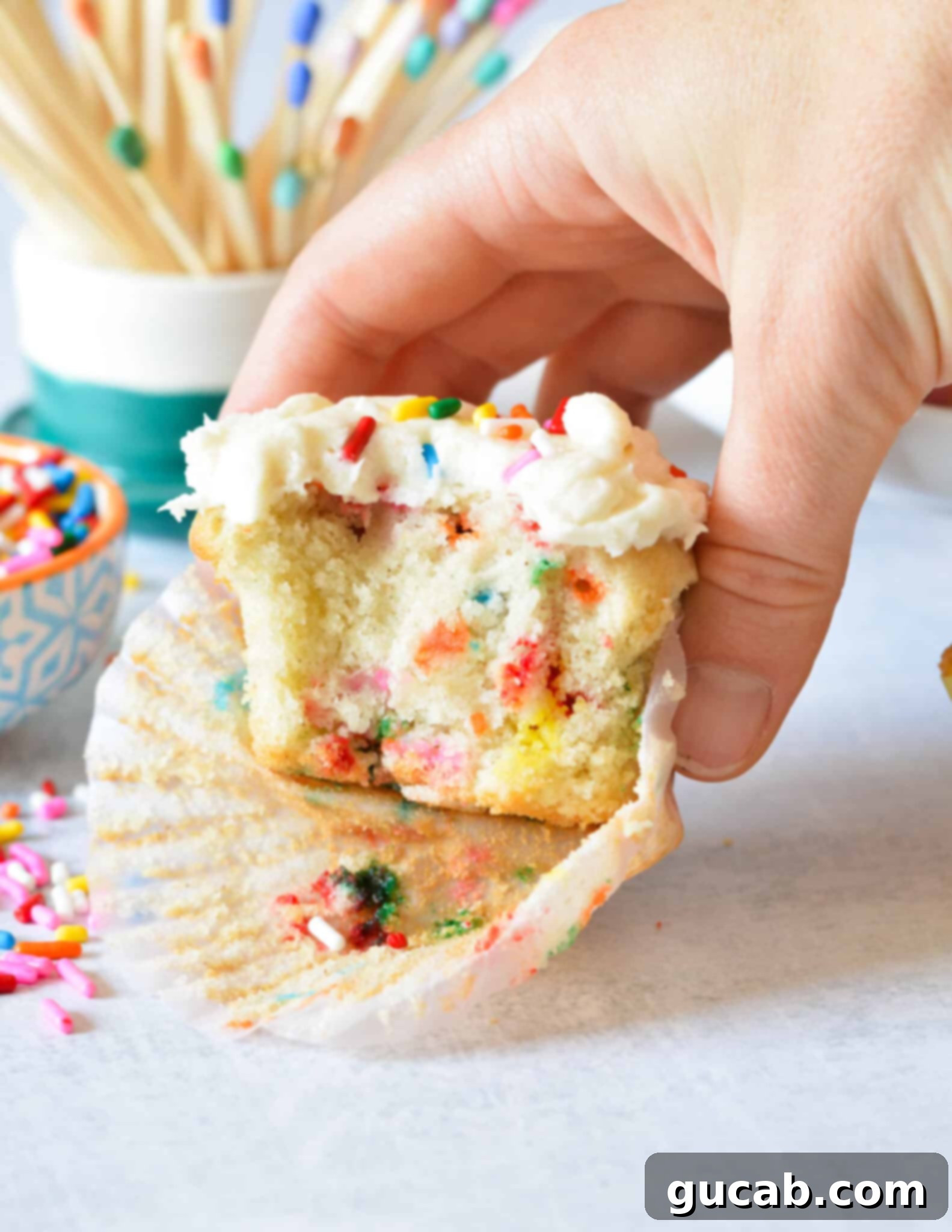 A hand holding a gluten-free funfetti cupcake with a bite taken out, revealing the colorful sprinkles inside.