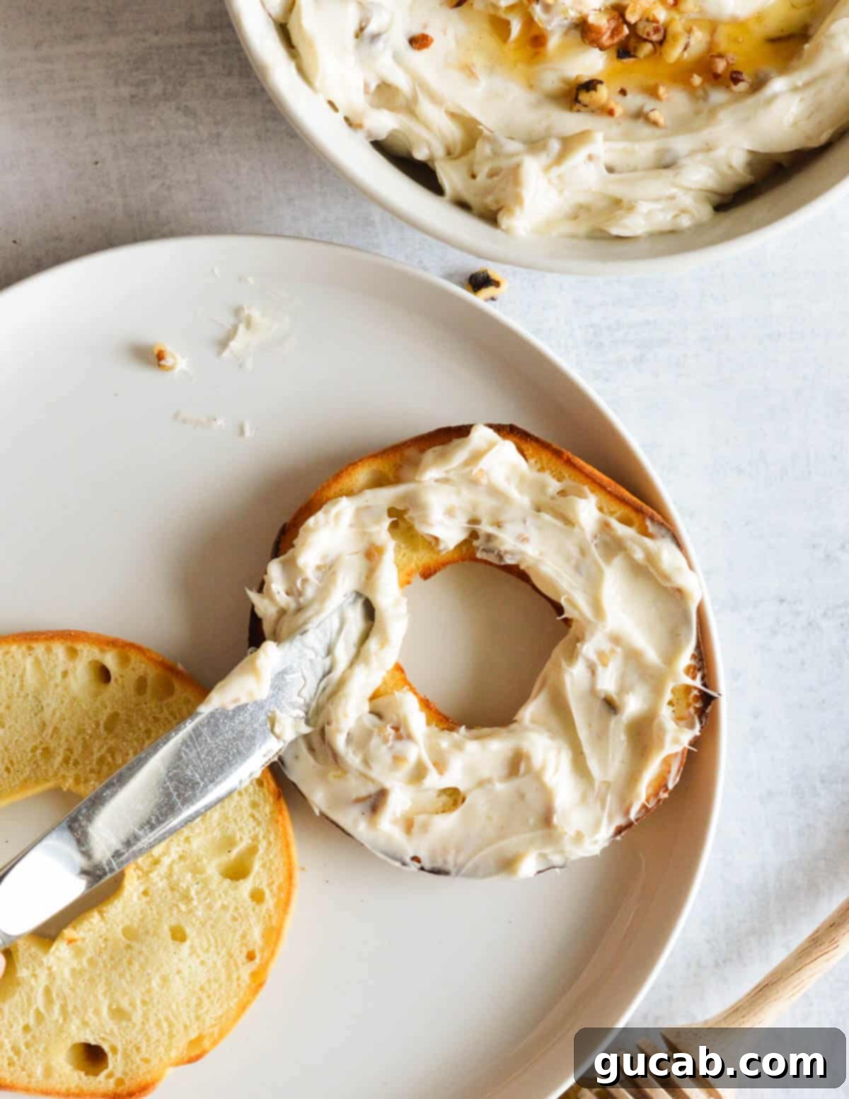 A hand spreading homemade honey walnut cream cheese generously on a toasted bagel half, highlighting its creamy texture and visible walnut pieces.