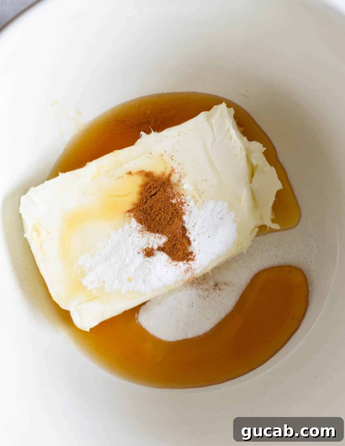 A large bowl containing a block of cream cheese, powdered sugar, ground cinnamon, and a pour of honey, ready to be mixed.