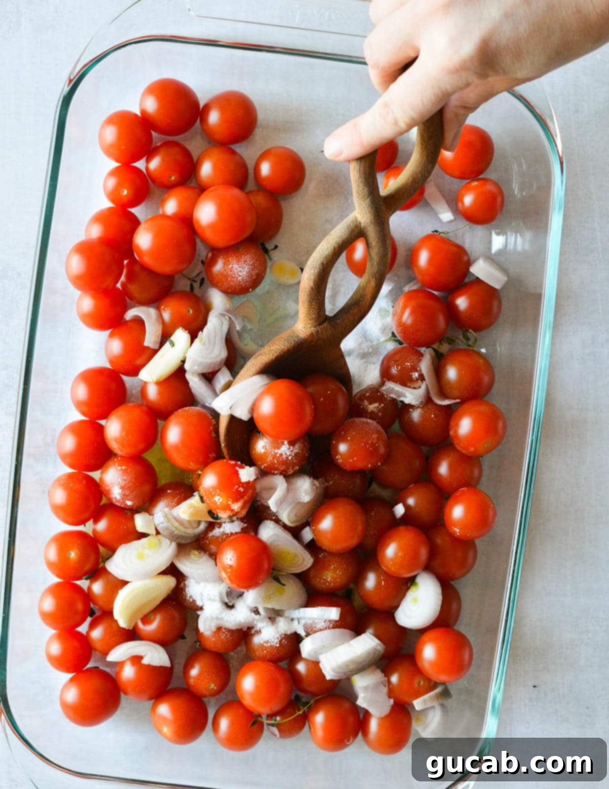 Cherry Tomato Soup 5 A hand with a wooden spoon gently stirring cherry tomatoes, olive oil, sliced shallots, and garlic in a baking dish, ensuring even coating before roasting.