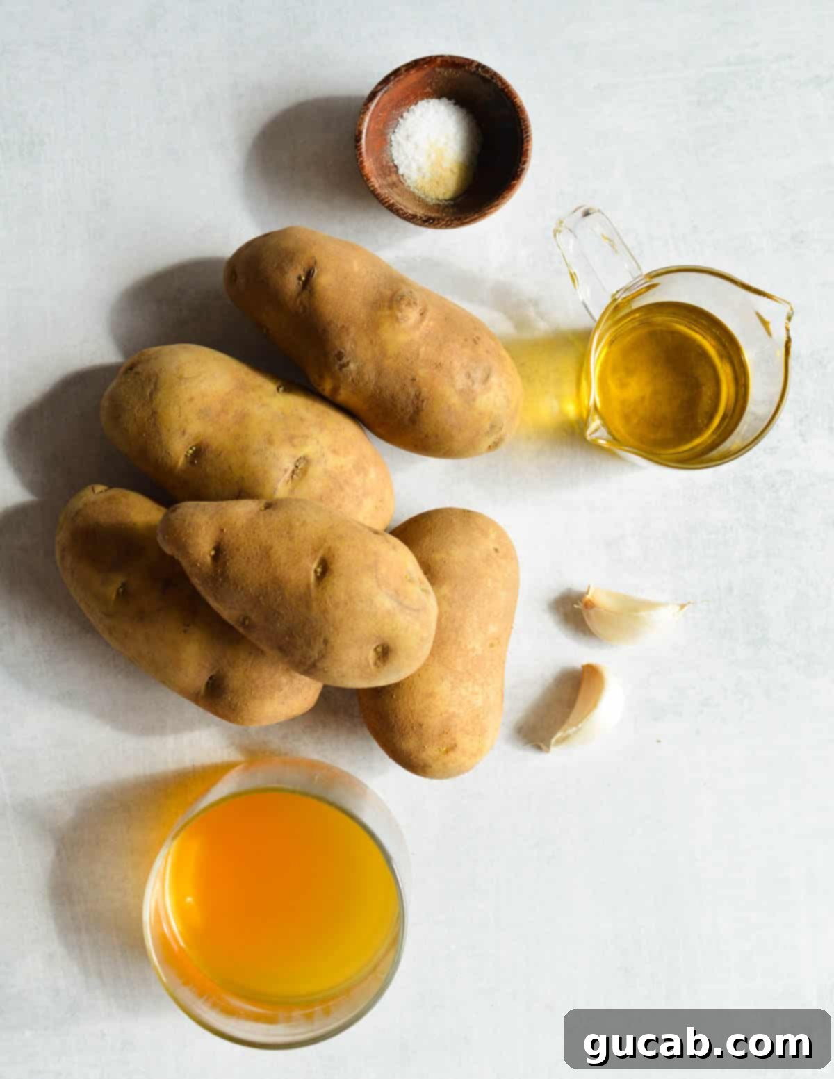 A selection of raw ingredients including russet potatoes, olive oil, salt, onion powder, garlic, and vegetable stock, laid out on a wooden surface.