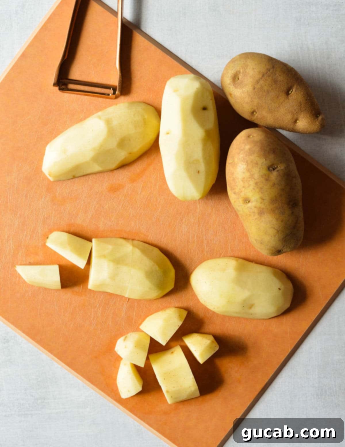 Russet potatoes on a cutting board, some whole and others cut into uniform chunks, ready for boiling.