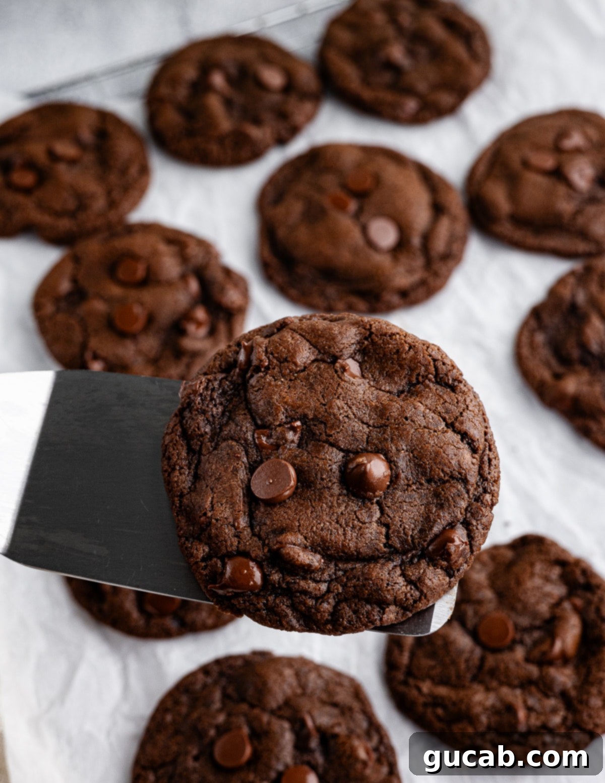 A spatula gently lifting a warm, freshly baked triple chocolate cookie from the baking sheet.