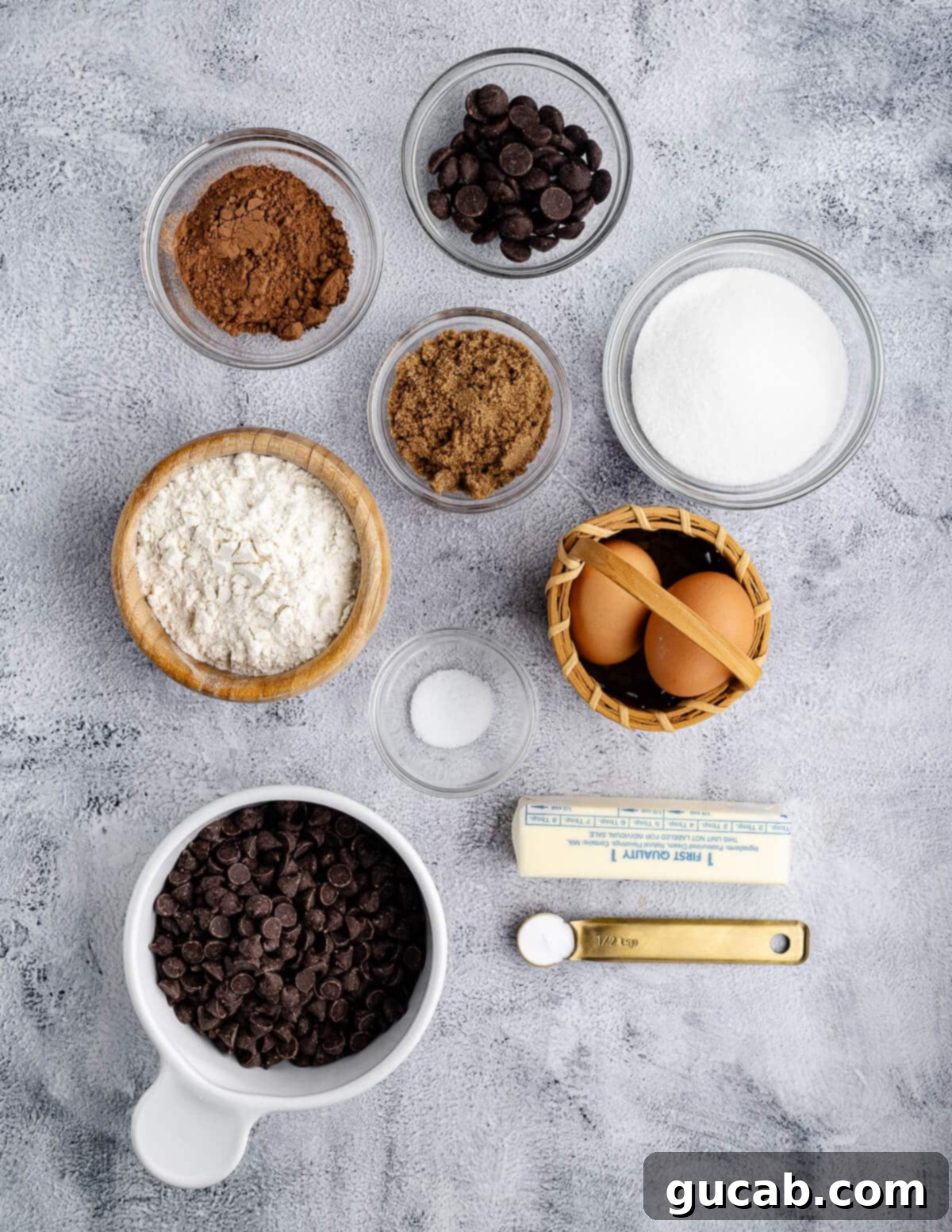 An array of triple chocolate cookie ingredients laid out: dark chocolate chips, brown sugar, granulated sugar, cocoa powder, eggs, all-purpose flour, salt, butter, and baking soda.