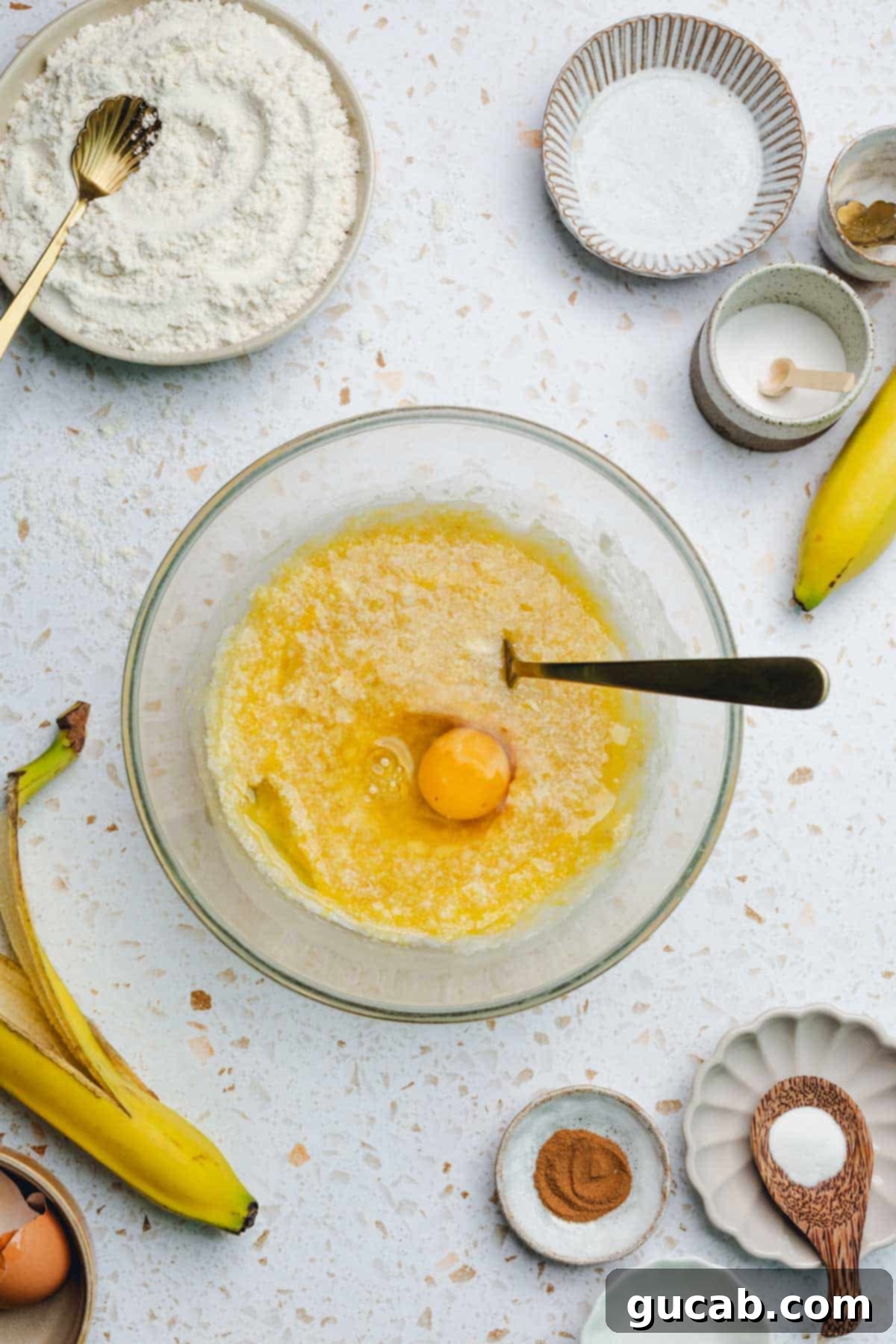A bowl of banana, oil, and honey mixture with a single egg cracked on top, ready to be mixed in.