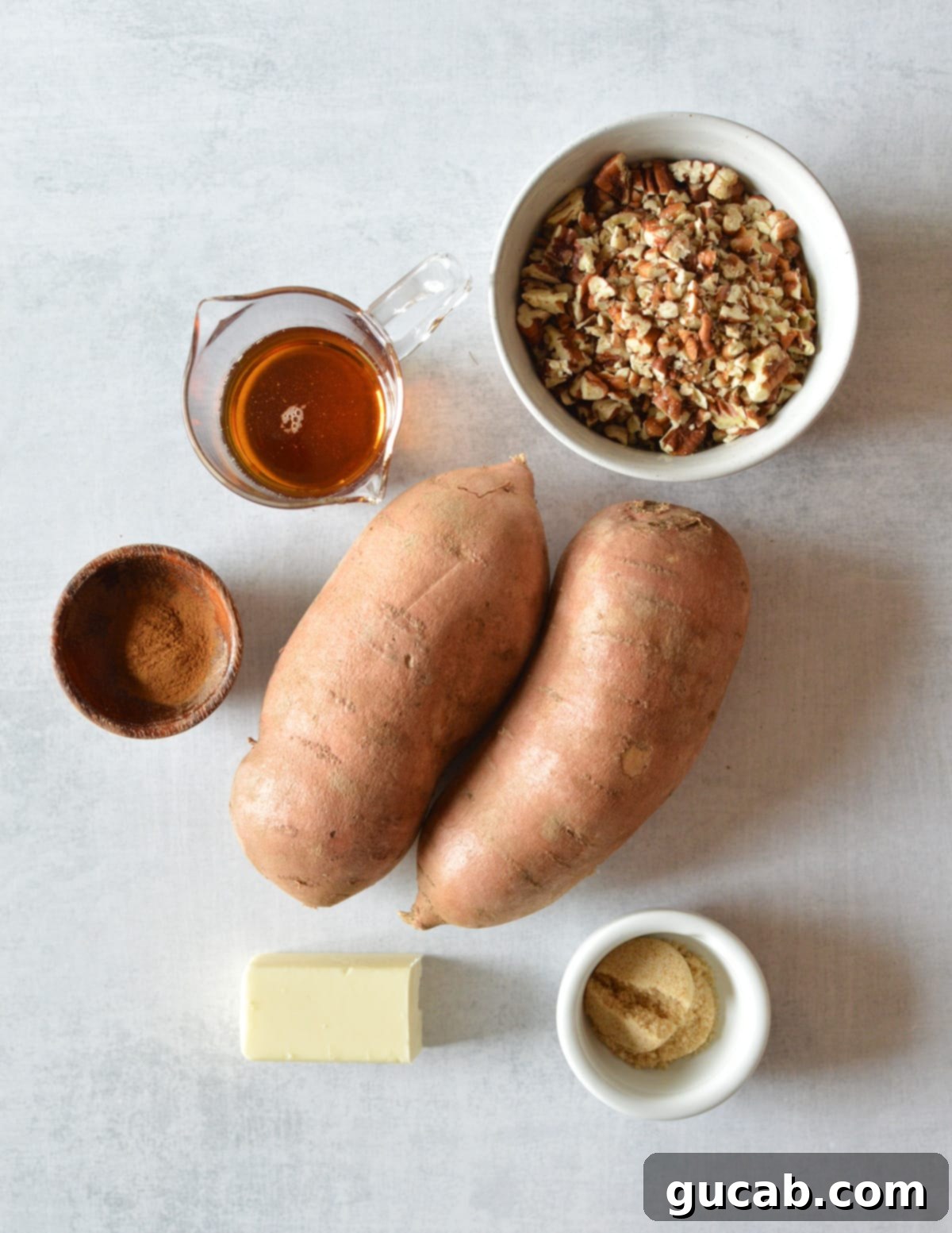 A collection of fresh ingredients: a bowl of chopped pecans, a bottle of maple syrup, a jar of cinnamon, raw sweet potatoes, butter, and brown sugar on a wooden surface.