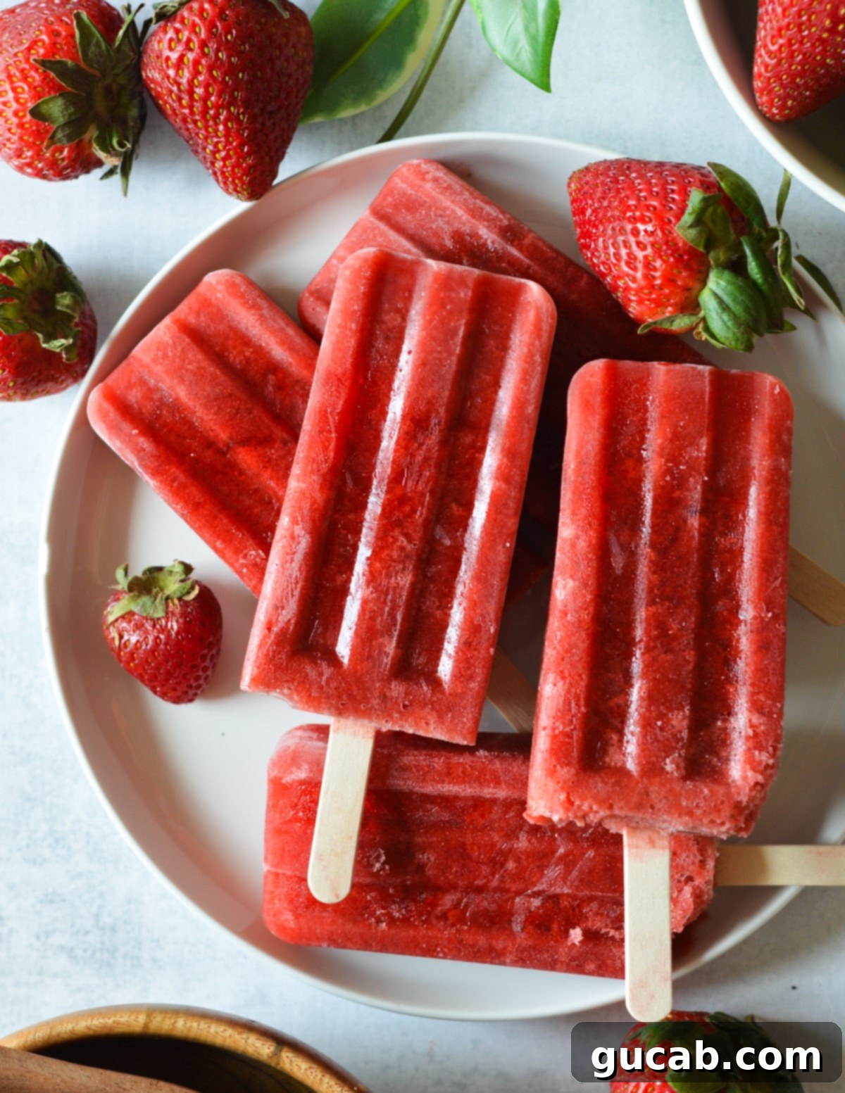 Vibrant homemade strawberry popsicles on a white plate, garnished with fresh, ripe strawberries, showcasing their natural red color and refreshing appeal.