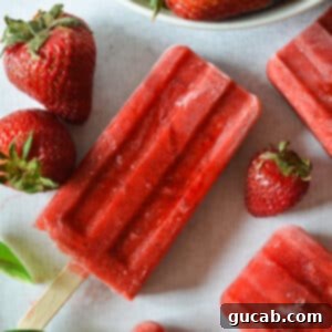 A single vibrant red strawberry popsicle, perfectly frozen, standing upright next to a scattering of fresh, ripe strawberries.