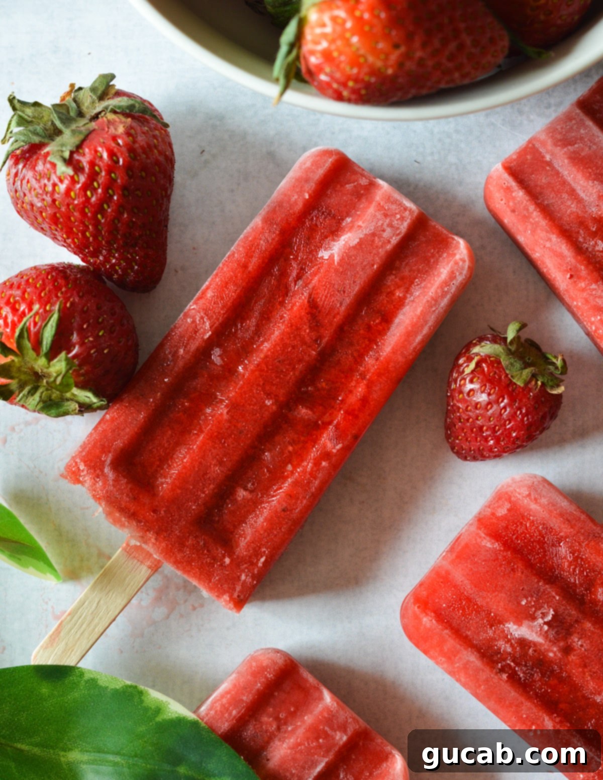 Several glistening homemade strawberry popsicles stand upright on a light-colored counter, surrounded by a scatter of fresh, vibrant strawberries, emphasizing their natural goodness.