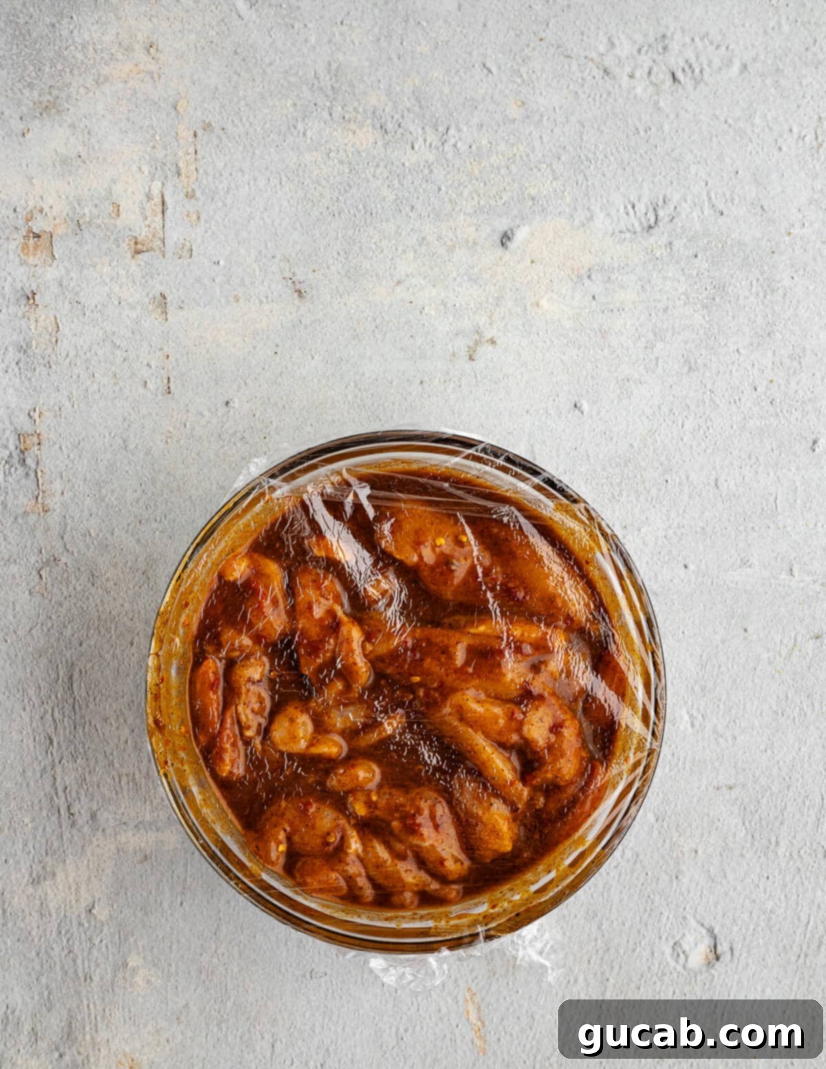 A bowl of chicken thighs fully coated in a chipotle marinade, covered with plastic wrap, marinating.